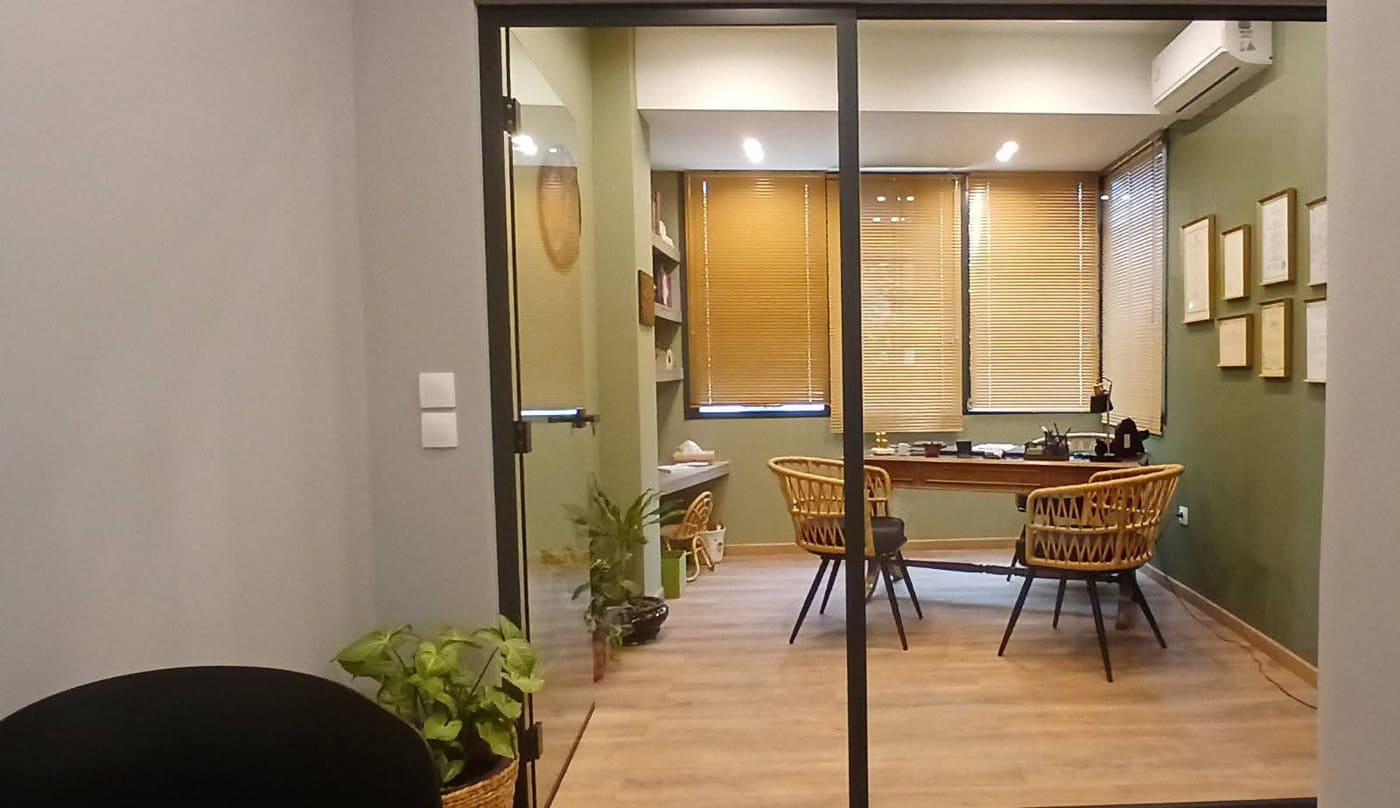 Το γραφείο όπως φαίνεται μέσα από τη γυάλινη πόρτα: πράσινος τοίχος, ξύλινα στόρια και οι δύο ψάθινες πολυθρόνες.